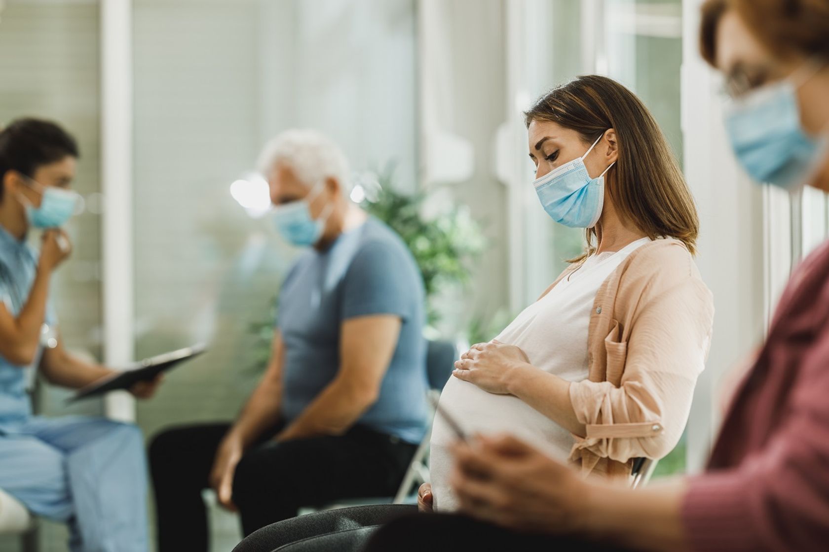 Schwangere Frau und andere Patienten sitzen mit Atemschutz in einem Wartebereich