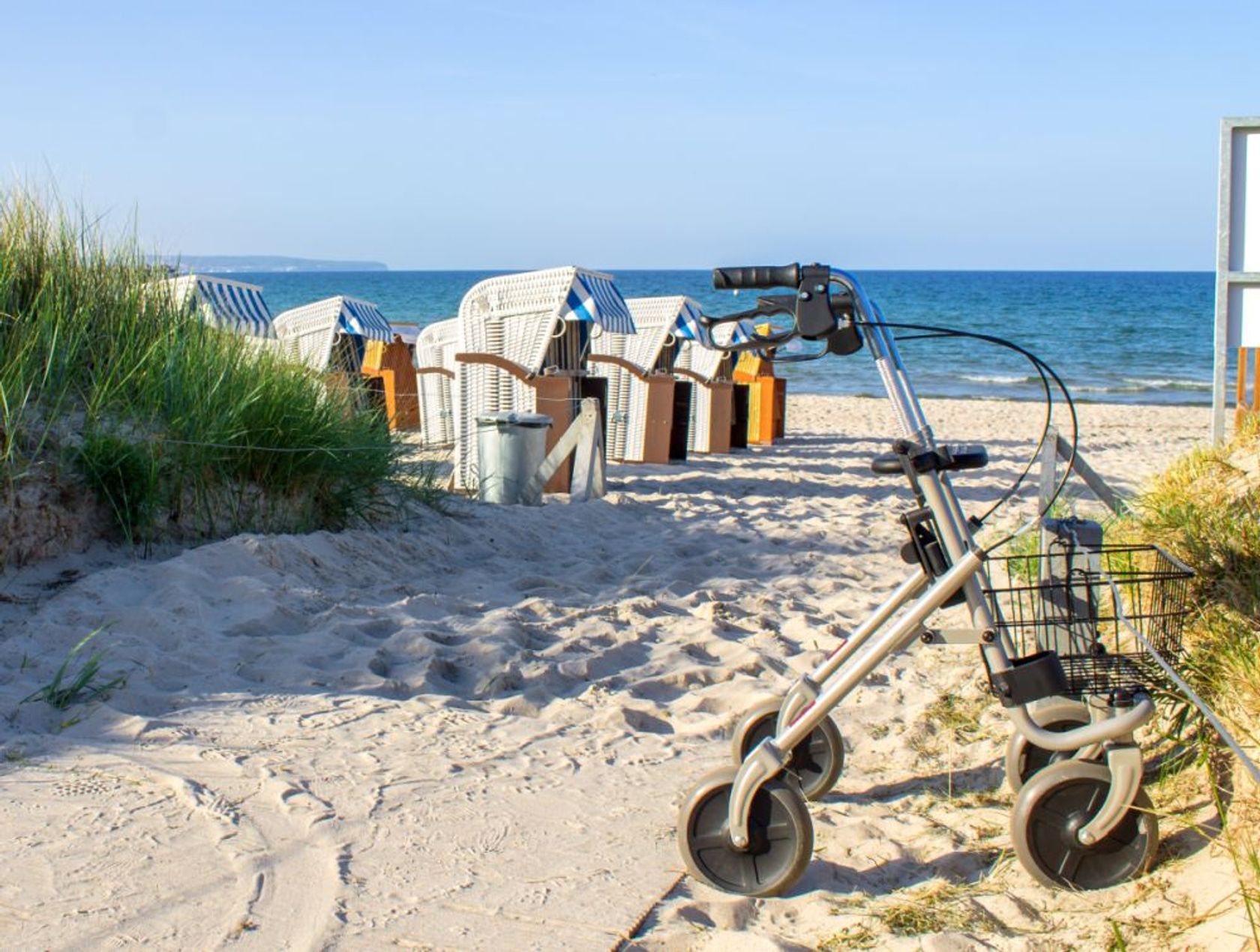 Ein Rollator ist an einem Strandübergang an der Düne geparkt, Blick auf Strandkörbe und die See.