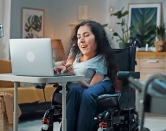 Kleinwüchsige Frau sitzt im Rollstuhl vor dem geöffneten Notebook