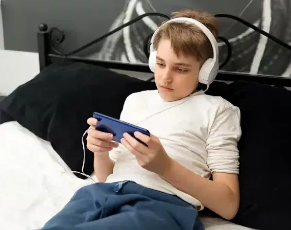 Müde aussehender Teenager-Junge mit Kopfhörern und Handy liegt auf dem Bett.