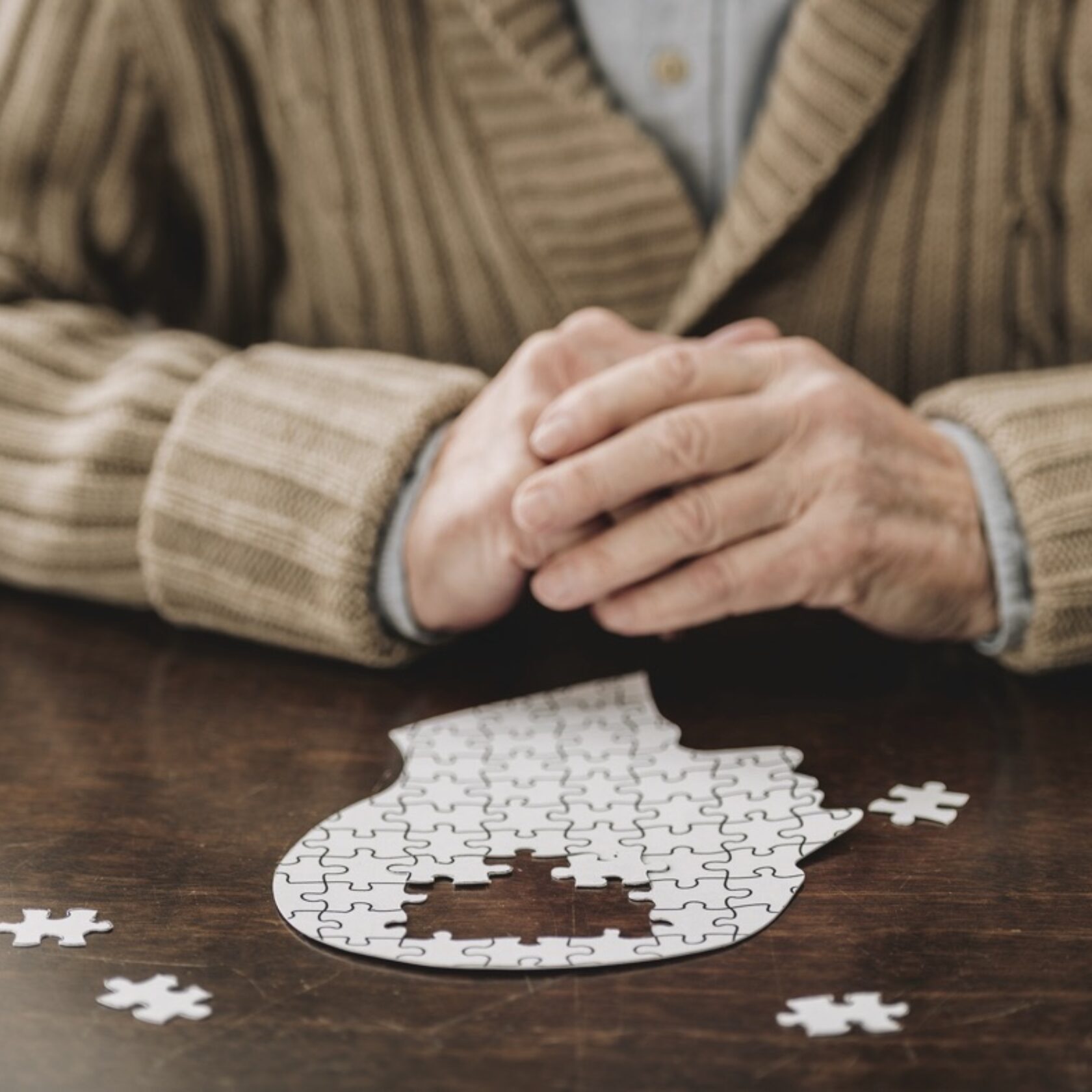 Senior sitzt mit gefalteten Händen vor dem Puzzle eines menschlichen Kopfes und hat Schwierigkeiten es zu vollenden