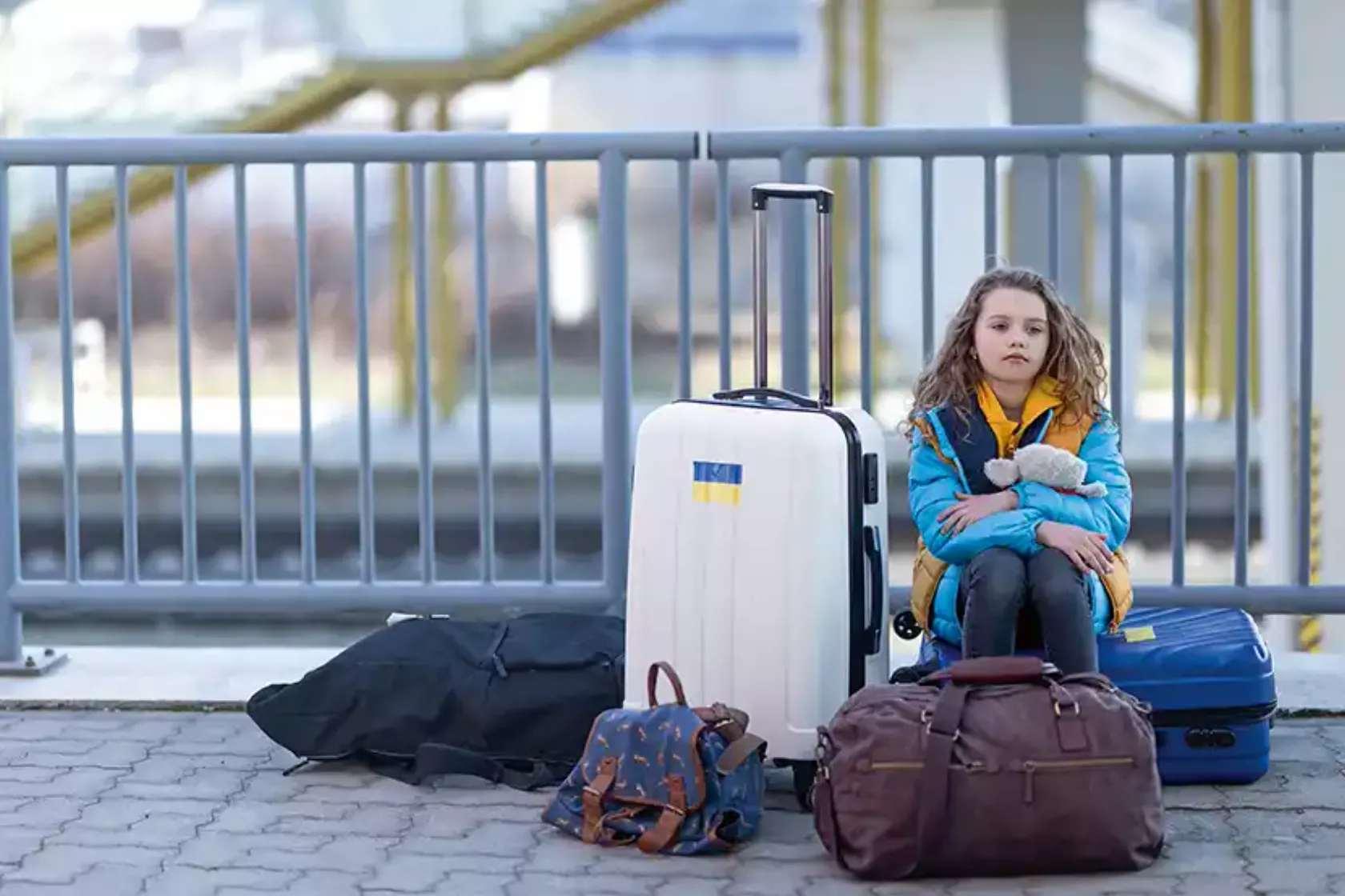 Mädchen in blauer Jacke mit gelbem Schal sitzt mit Gepäck auf dem Boden am Bahnhof.