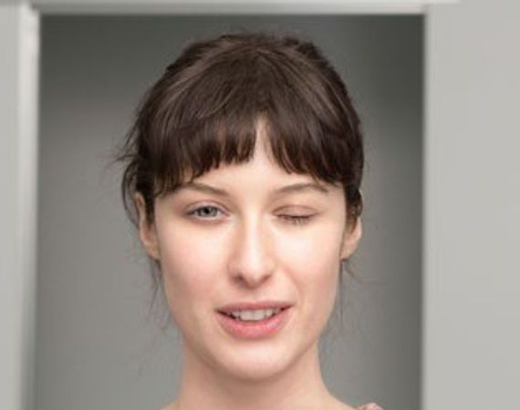 Porträt einer jungen Frau, deren linkes Auge geschlossen ist, während sie mit dem rechten geradeaus blickt.
