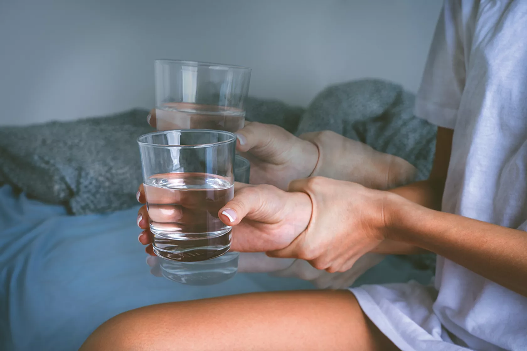 Foto einer Hand, die zittert - die Person hält ein Wasserglas und hält mit der anderen Hand das Handgelenk fest.